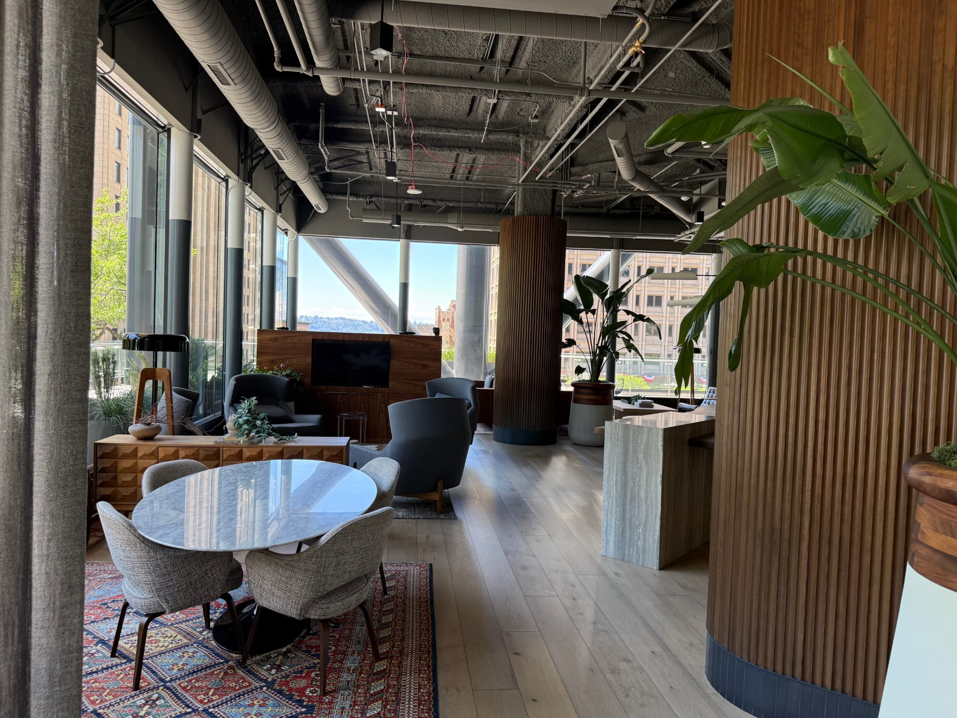 Tenant lounge interior with a round marble table, upholstered chairs, wood columns, and floor-to-ceiling windows overlooking the city.
