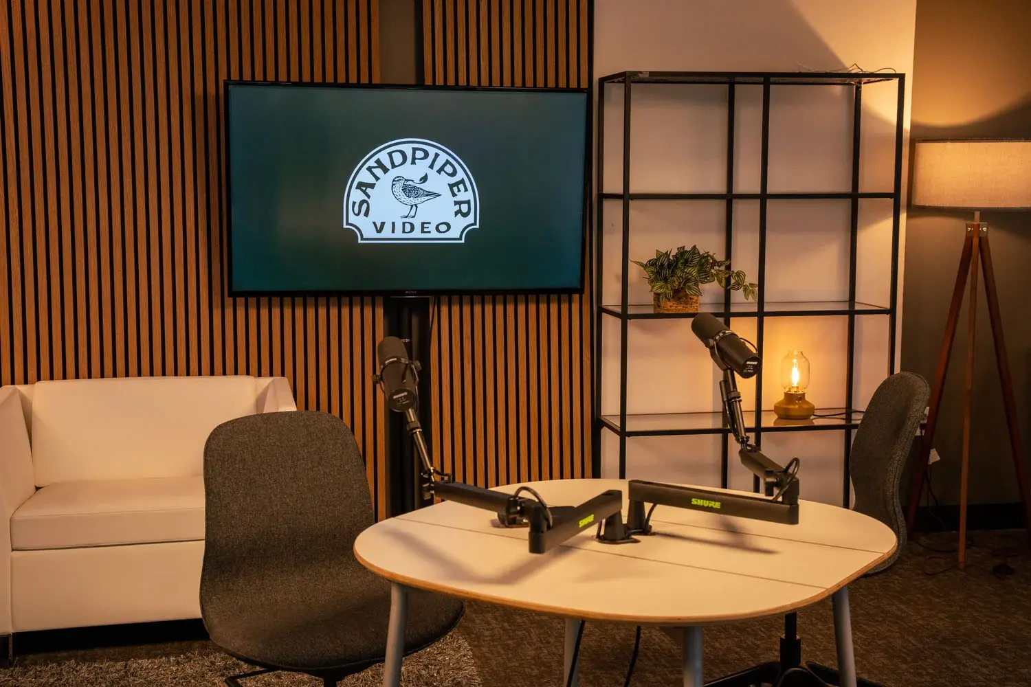 Sandpiper Video podcast set at SURF with a wood-slat backdrop, Shure microphones on boom arms, a white sofa, and warm lamp lighting.