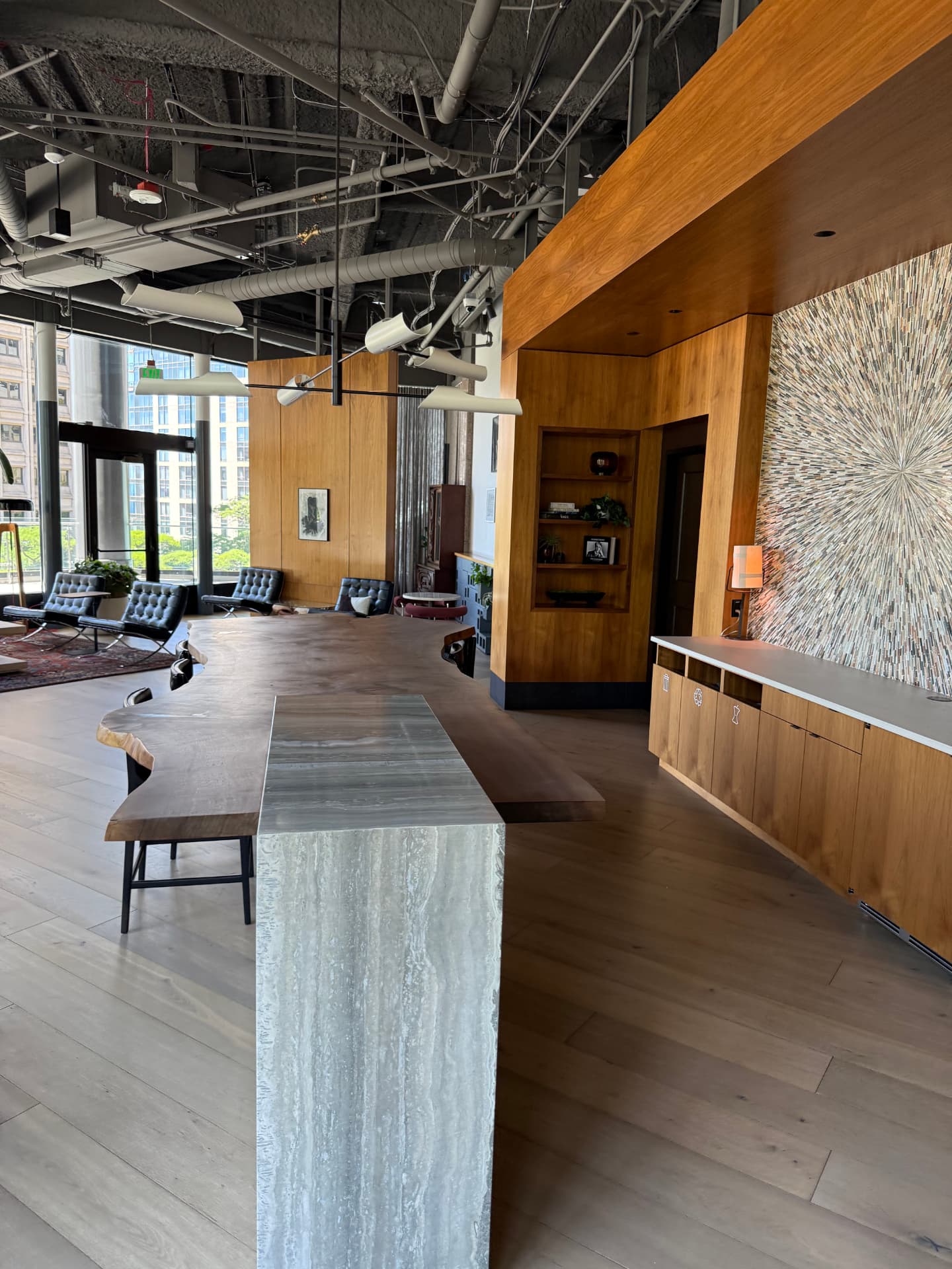 Long live-edge wood communal table with a travertine base, next to a wall of black leather Barcelona chairs.