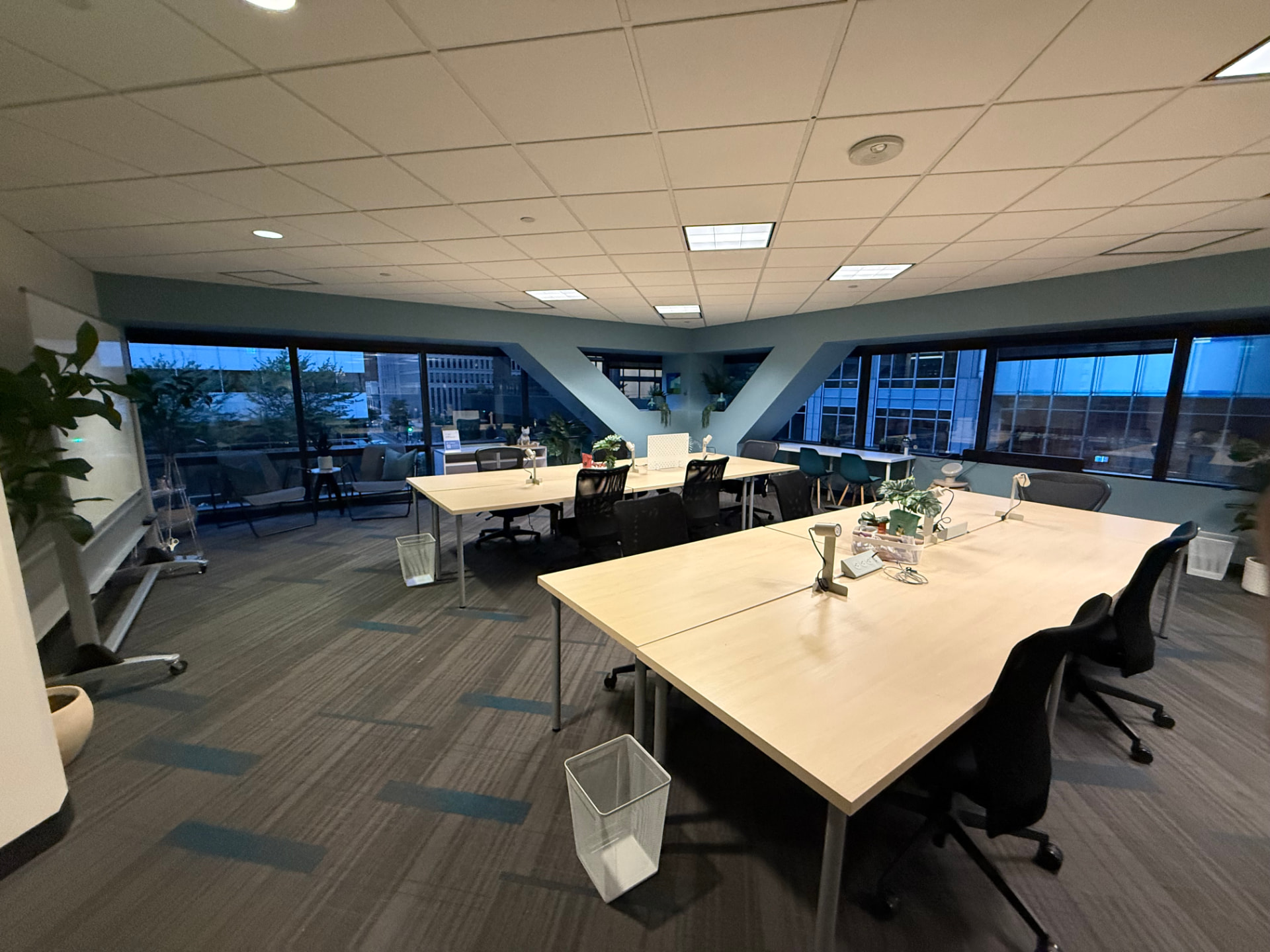 Main coworking area at The SWELL with two long light-wood tables, black mesh chairs, and a downtown Seattle view at dusk.